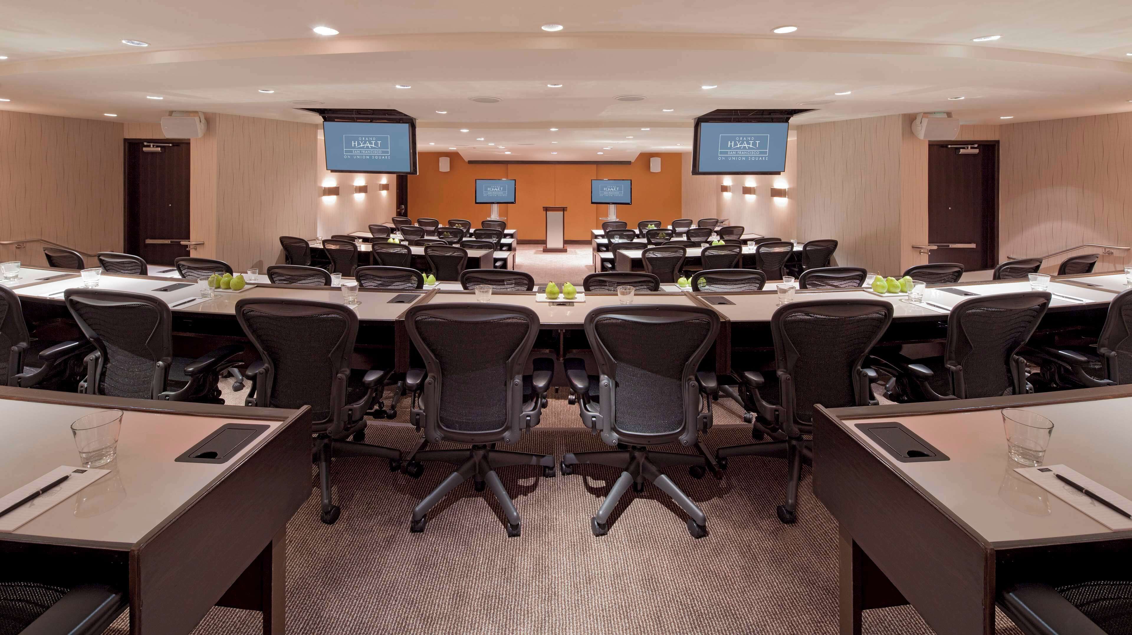 Un salón de conferencias con varias mesas y sillas, equipado con pantallas y un atril.
