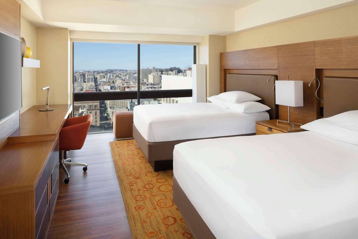 A hotel room with two beds, a desk, a chair, and a large window offering a city view.