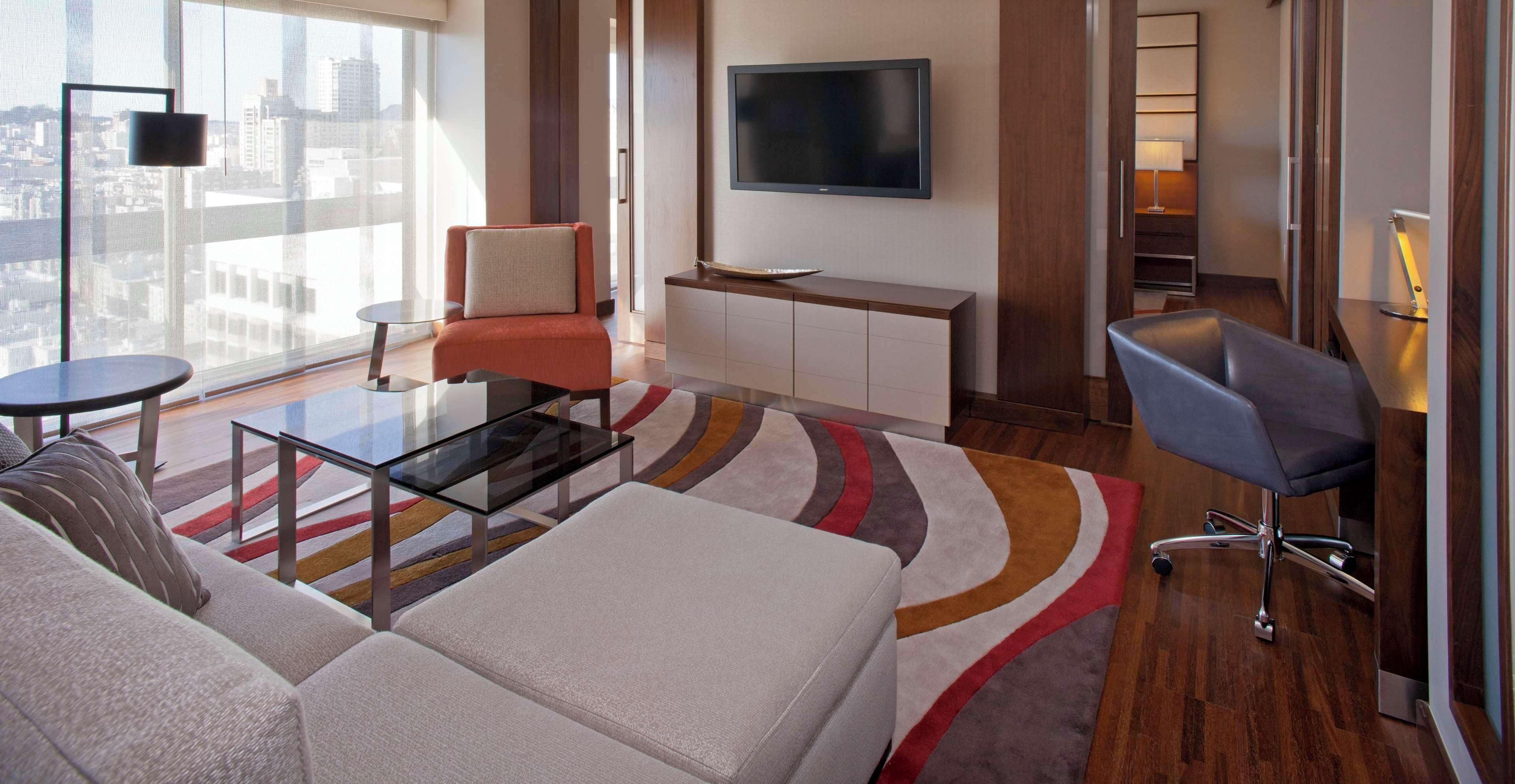 A modern hotel room with a large window, a flat-screen TV, a sofa, a coffee table, a chair, and a desk with a lamp.