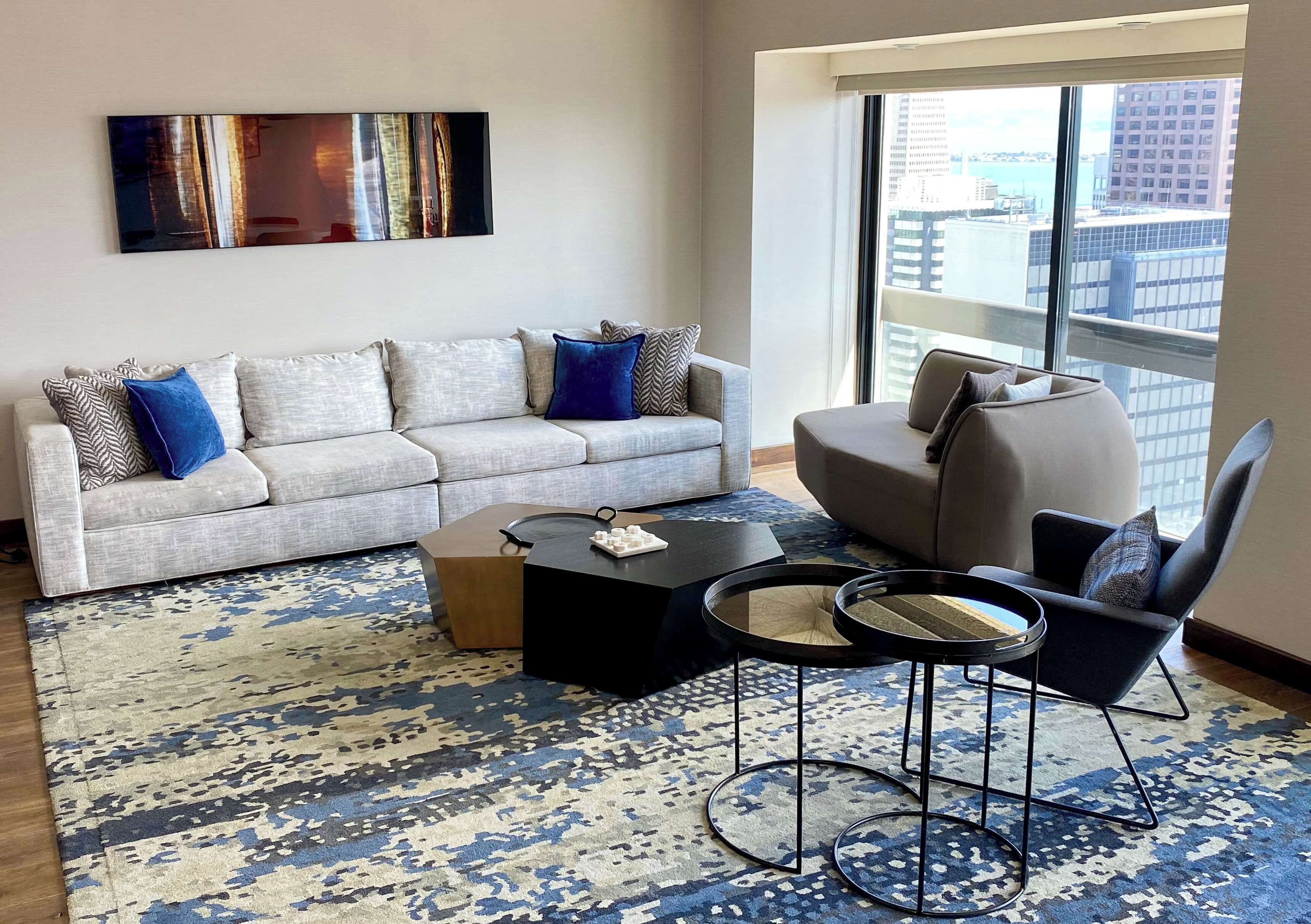 Un salon moderne avec un canapé gris, un tapis aux motifs bleus et blancs, et une vue sur la ville à travers de grandes fenêtres.