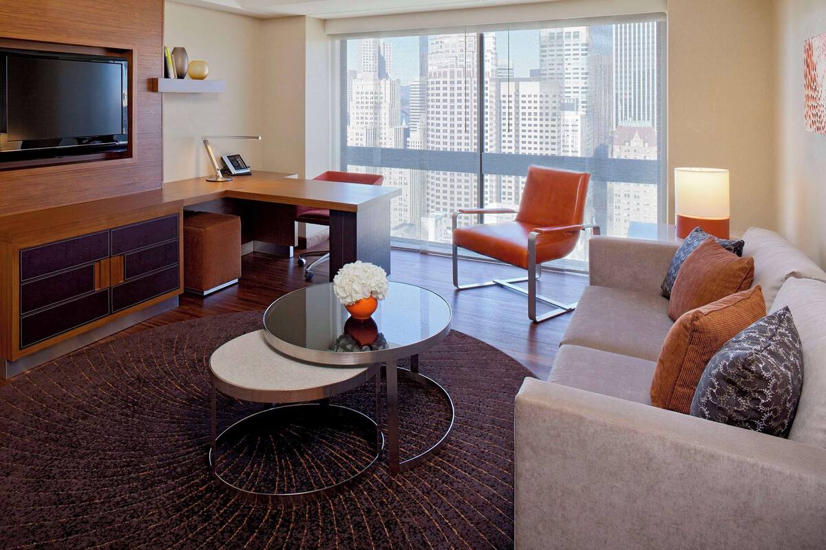 A modern living room with a flat-screen TV, a desk, a round coffee table, and a sofa with decorative pillows.