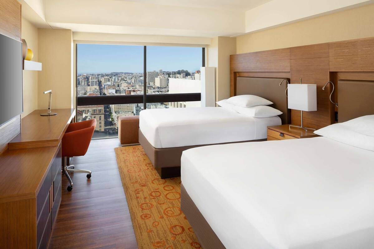 A hotel room with two beds, a desk, a chair, a large window with a city view, and a wooden headboard.