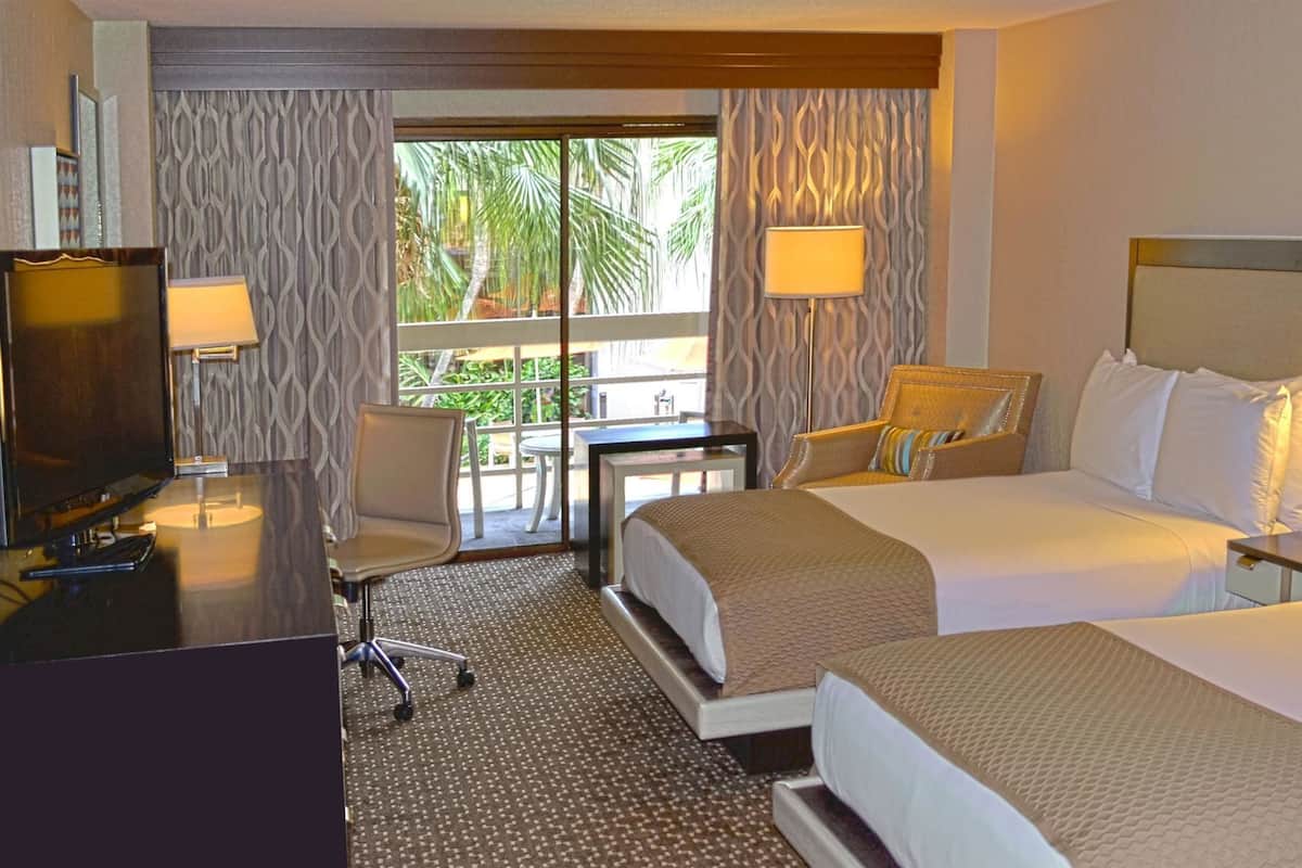 Une chambre d’hôtel avec deux lits, un bureau, une chaise et un balcon donnant sur des palmiers.