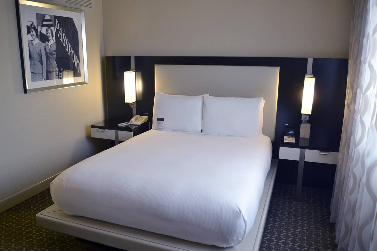 A hotel room with a large bed, two bedside tables, a nightstand, a framed picture, and a wall-mounted clock.