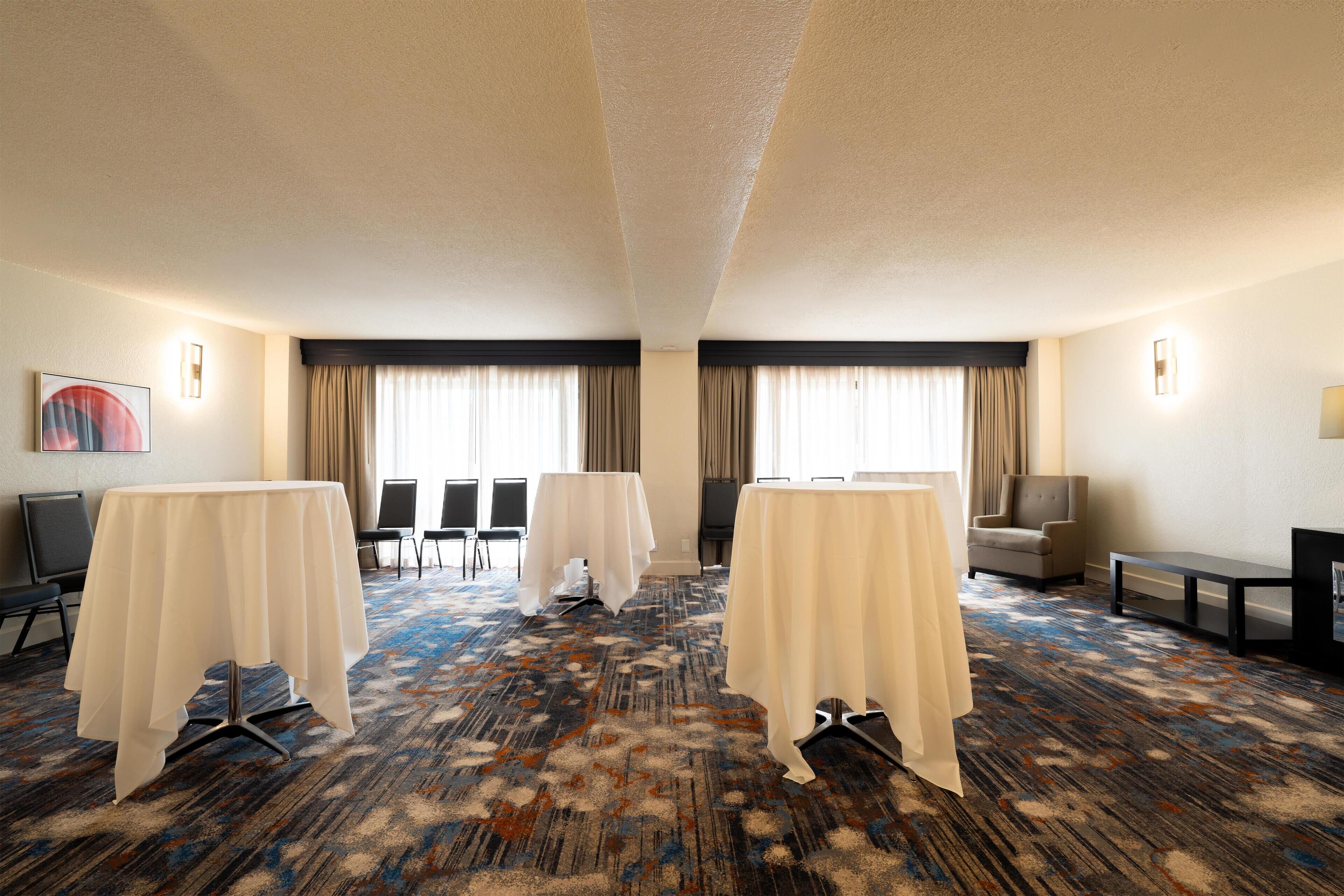 A hotel room with a carpeted floor, two tables with white tablecloths, a chair, a sofa, a TV, and a painting on the wall.