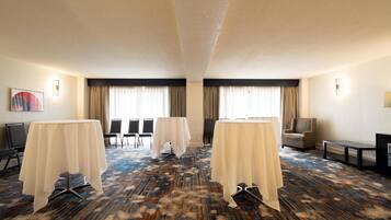 A hotel room with a carpeted floor, two tables with white tablecloths, a chair, a sofa, a TV, and a painting on the wall.