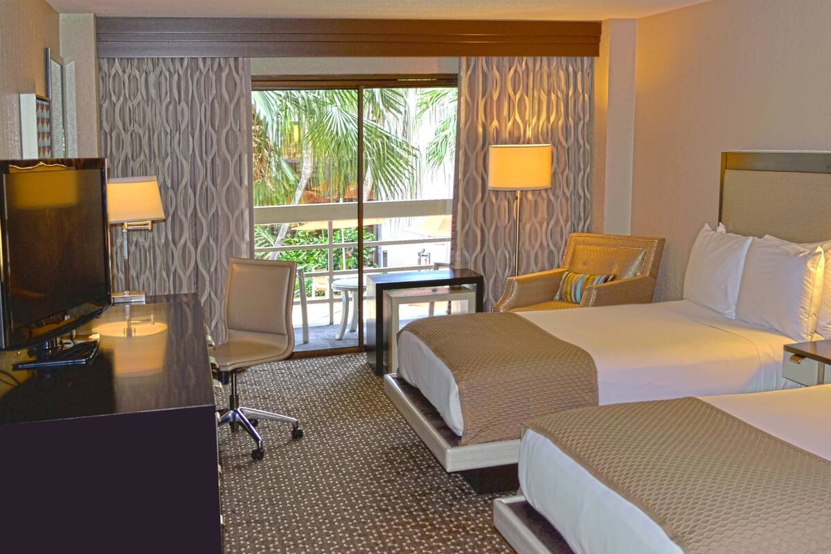 A hotel room with two beds, a desk, a chair, and a balcony with a view of palm trees.