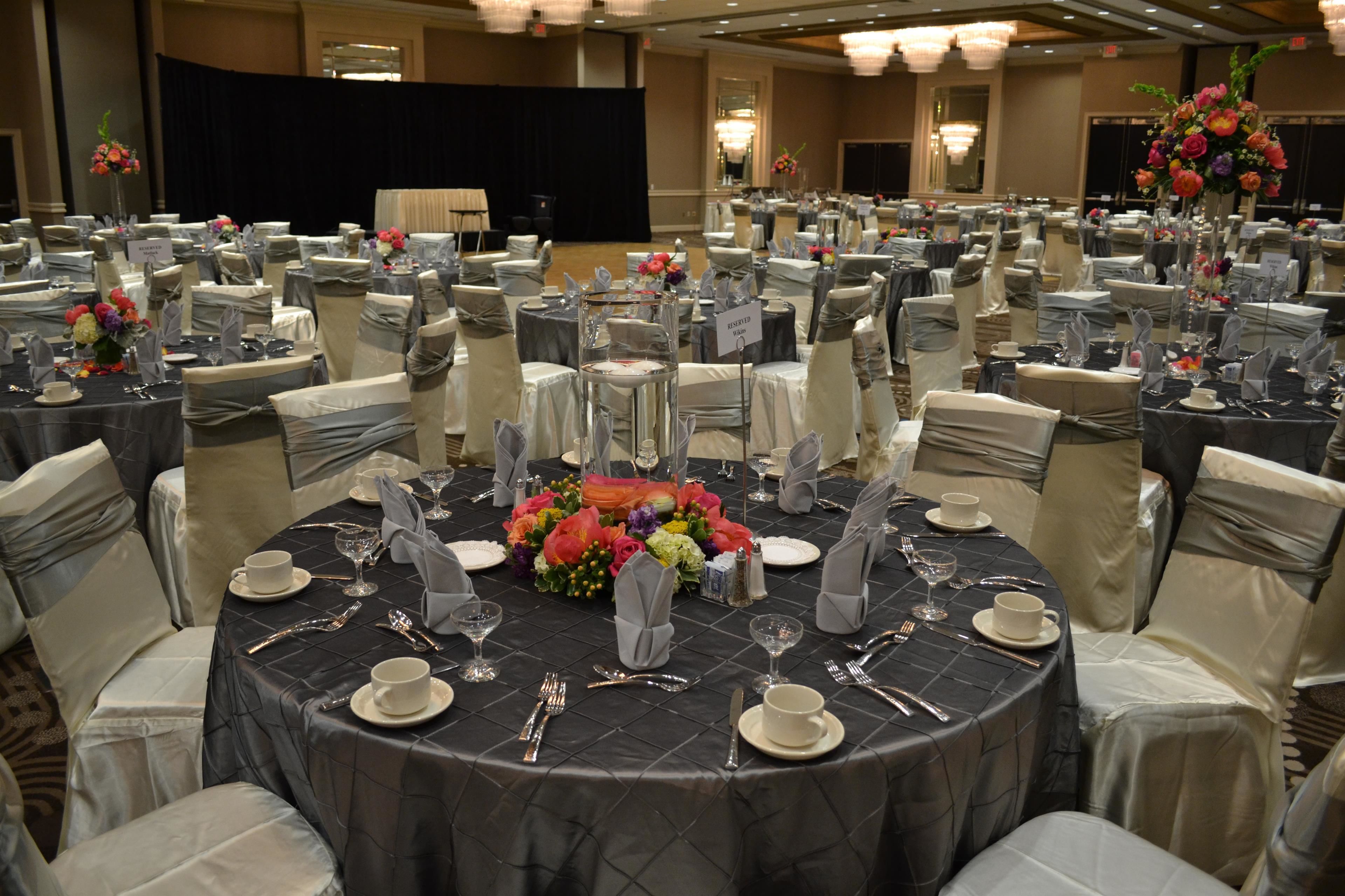 Un salón de banquetes con mesas redondas dispuestas para un evento formal, cada una con loza, cubiertos y centros de mesa florales.