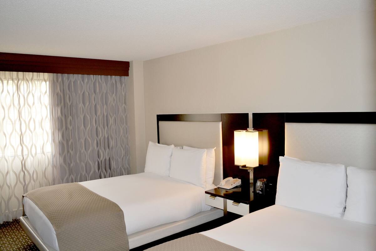 A hotel room with two beds, a bedside table with a lamp, and a window with patterned curtains.