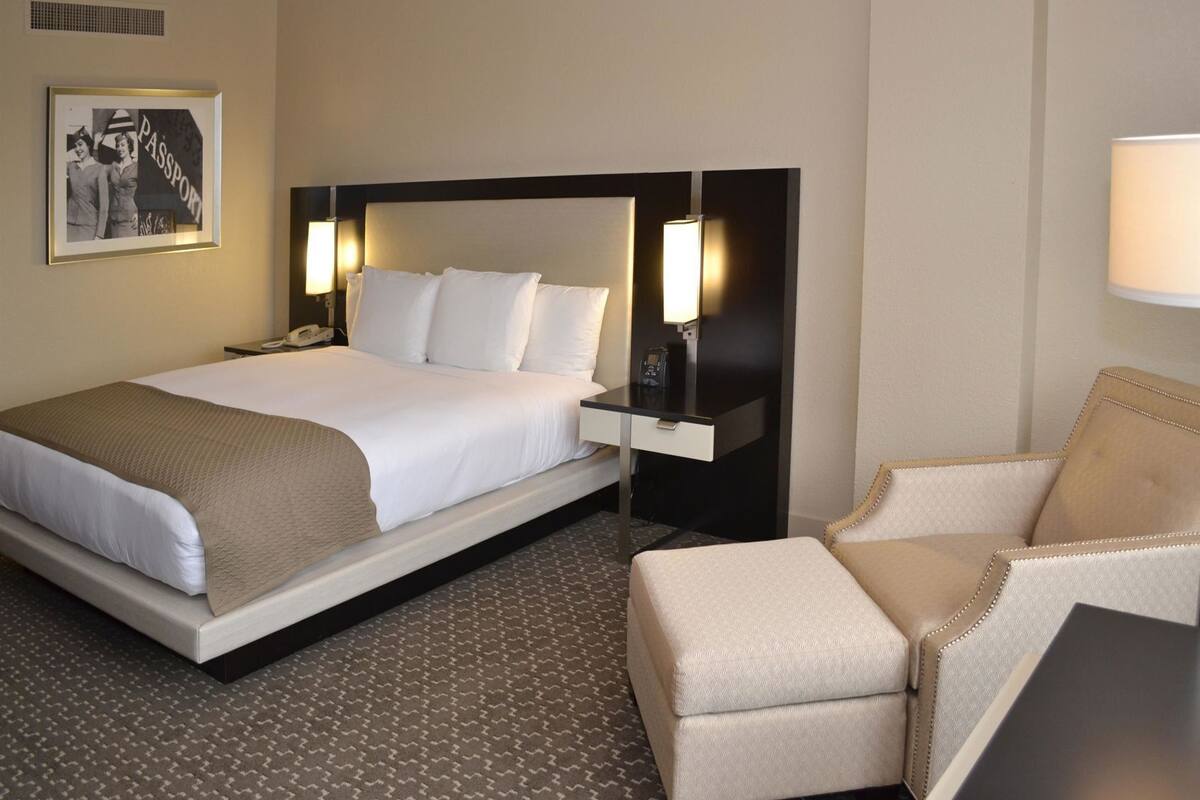 Une chambre d’hôtel avec un lit, des tables de chevet, une chaise, une lampe et un tableau encadré au mur.