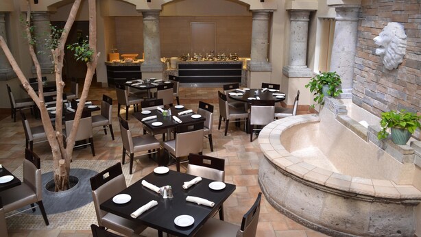 An indoor dining area with tables and chairs, a central fountain, and a stone wall with a lion sculpture. - Doubletree by Hilton Houston Hobby Airport (Houston)