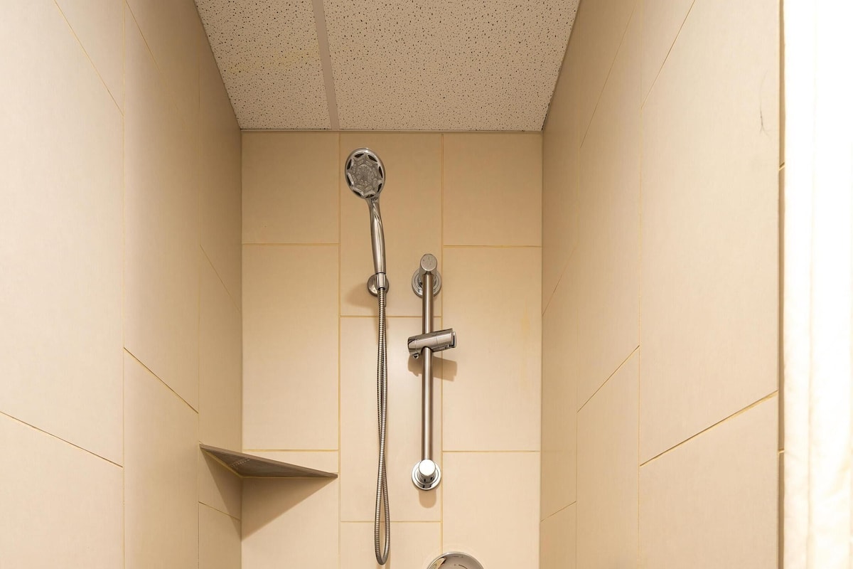 A small, tiled shower stall with a handheld showerhead, a grab bar, and a safety bar on the floor.