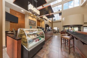 Una cafetería moderna con un expositor de vidrio lleno de pasteles y bebidas, una máquina de café y una zona de estar con taburetes de bar.