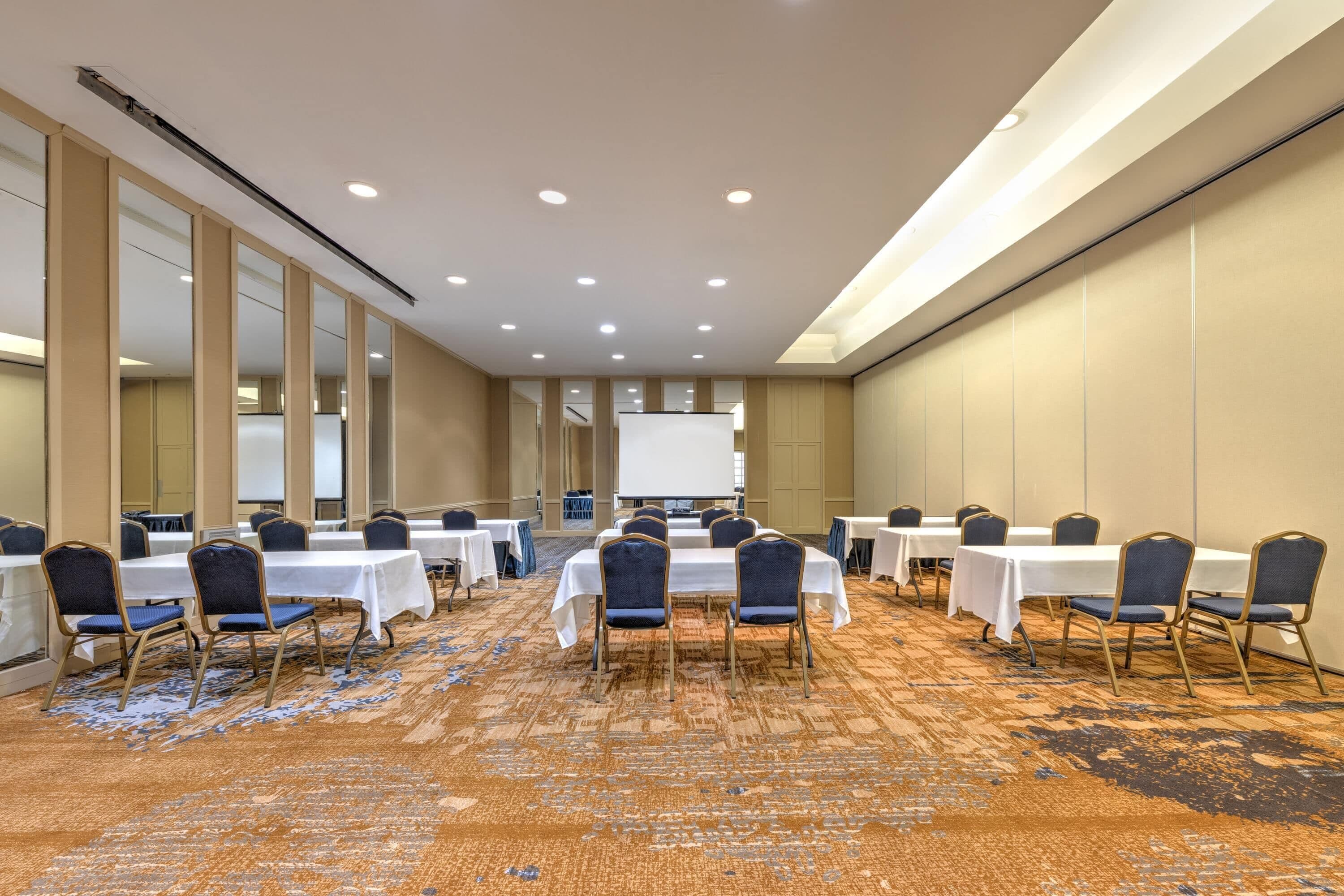 A conference room with multiple tables and chairs, a large screen at the front, and a carpeted floor.