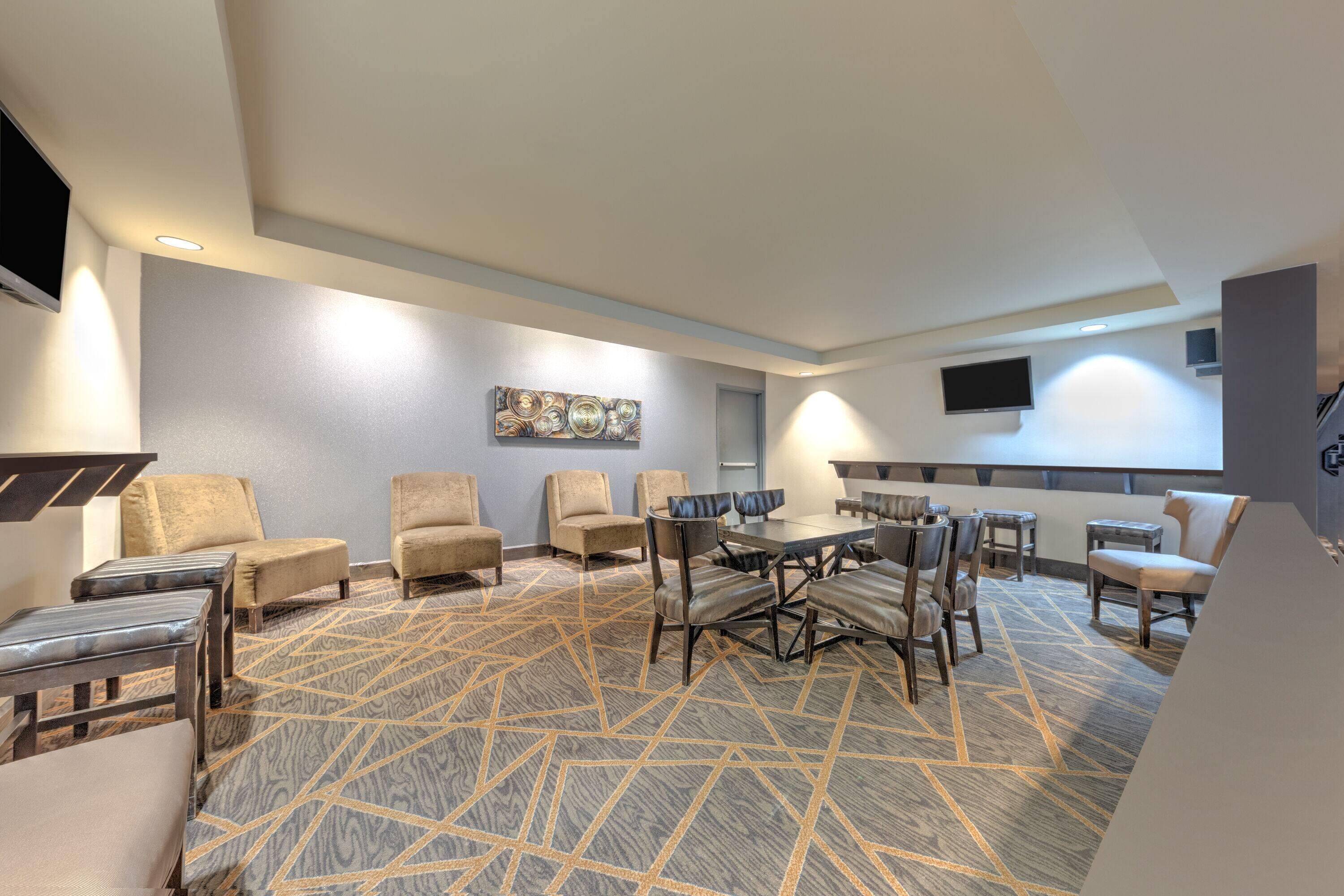 A hotel lobby with a geometric patterned carpet, multiple seating areas with sofas and chairs, a central dining table, and a flat-screen TV mounted on the wall.