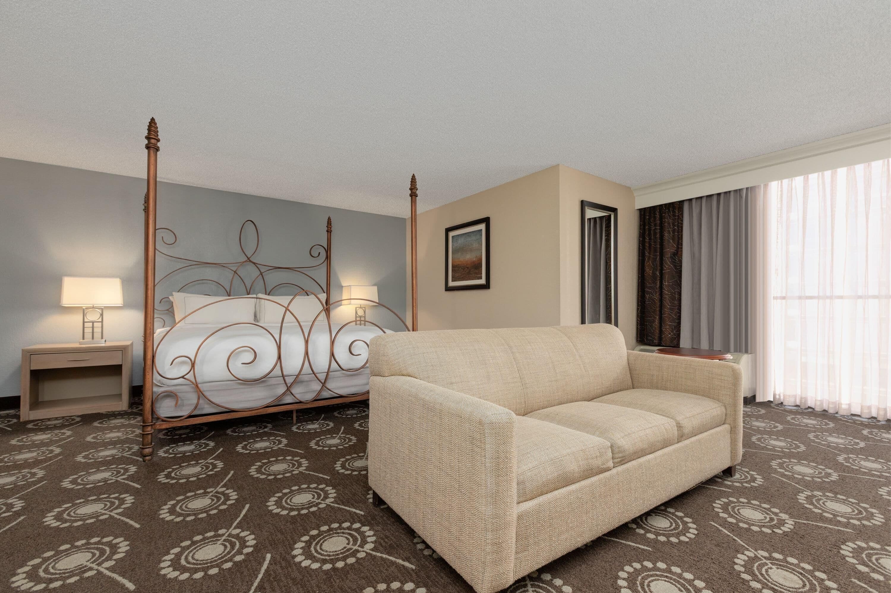 Habitación de hotel con cama, sofá, mesita de noche, lámpara y ventana con cortinas.