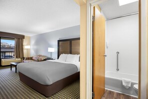 A hotel room with a large bed, a bedside table, a lamp, a window with curtains, and a bathroom with a bathtub and toilet.