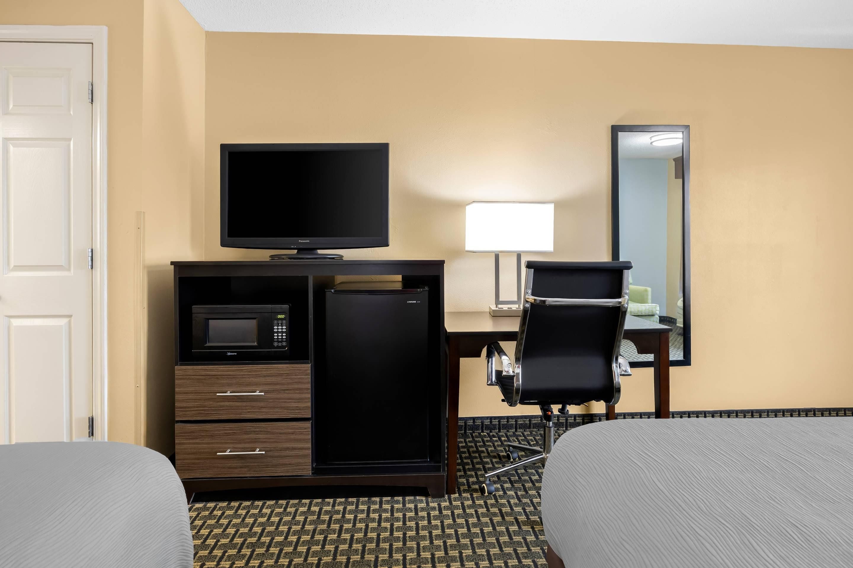 A hotel room with a bed, a desk with a television, a microwave, a refrigerator, a chair, and a mirror.