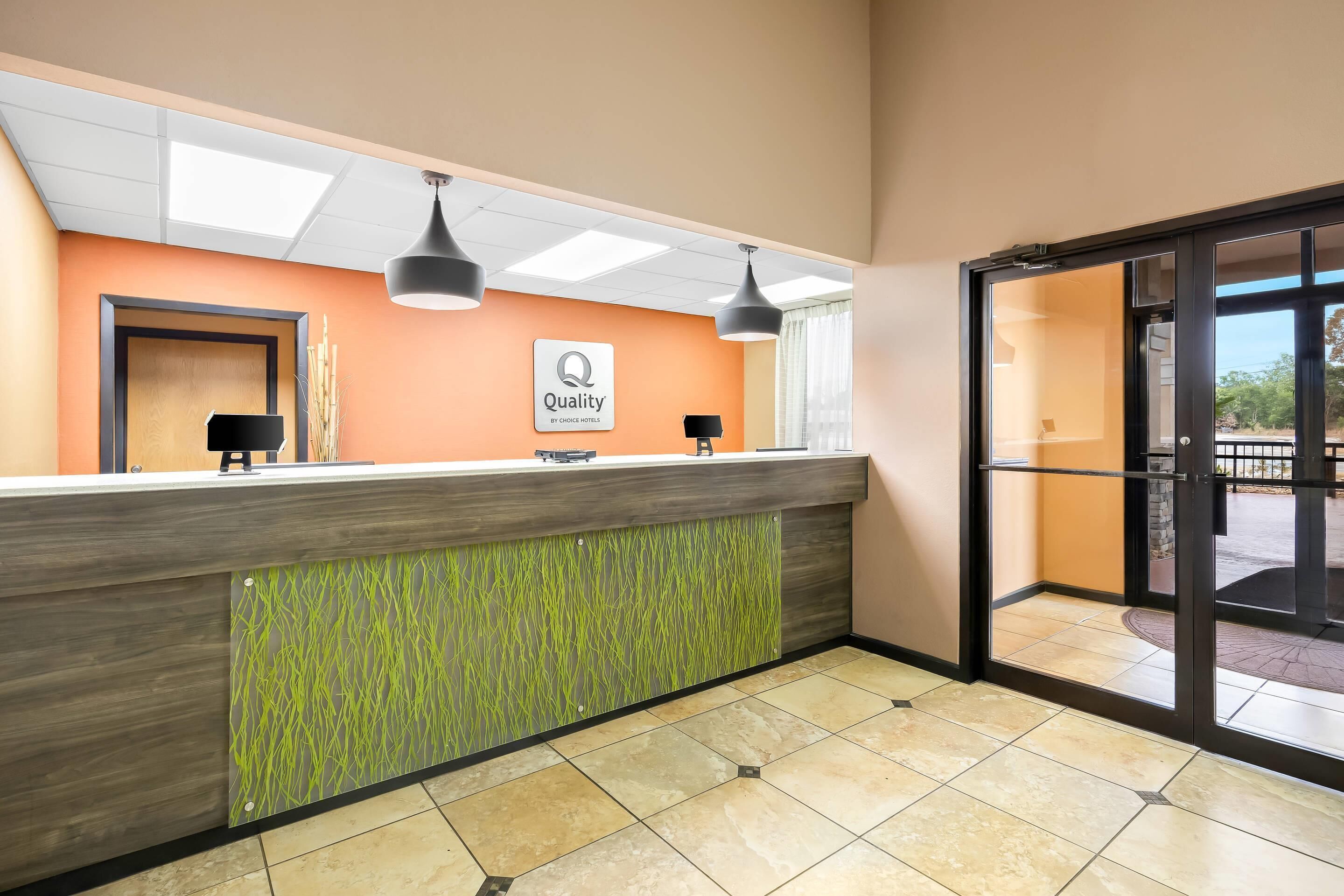 A hotel reception area with a wooden reception desk, a green patterned wall, and a sign indicating 'Quality by Chase Hotel'.