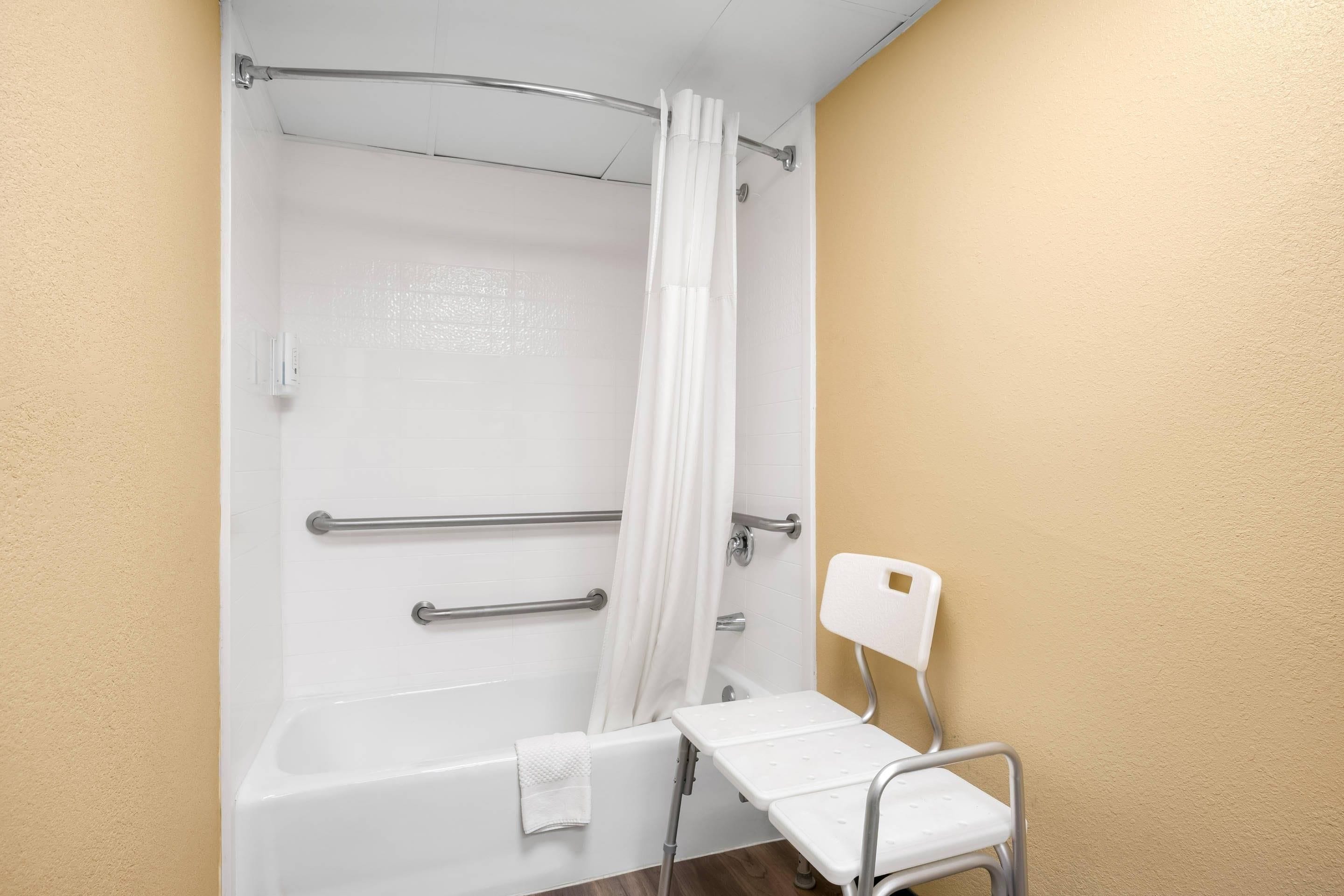 Un baño con bañera, cortina de ducha, una silla blanca y barras de apoyo.