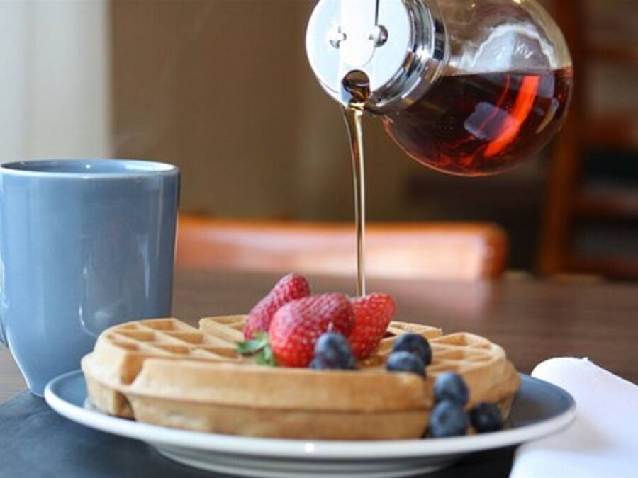 Un gofre con fresas y arándanos en un plato, una taza azul y jarabe siendo vertido sobre el gofre.