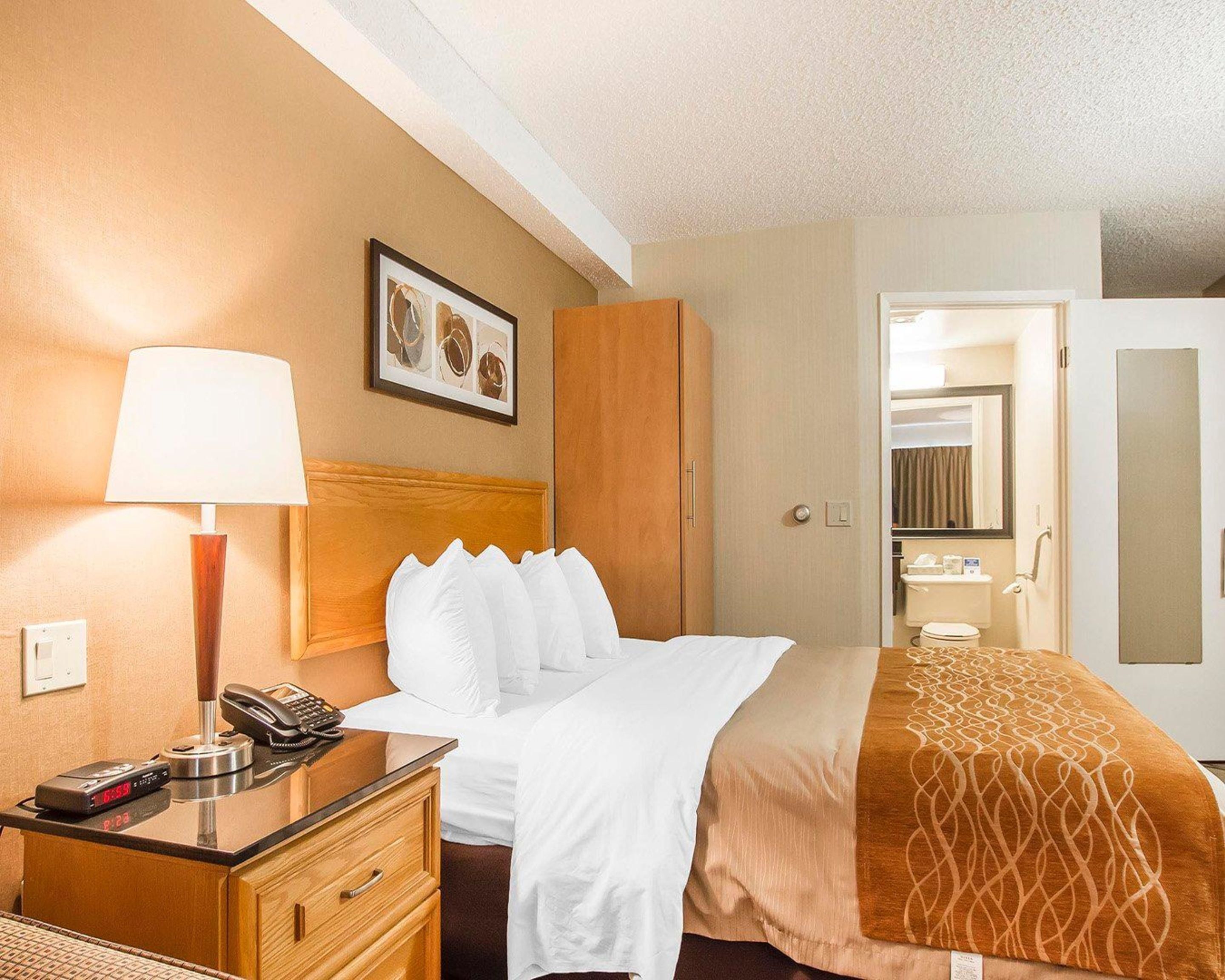 Habitación de hotel con cama, mesita de noche con lámpara, teléfono, reloj, armario y baño visible a través de una puerta abierta.
