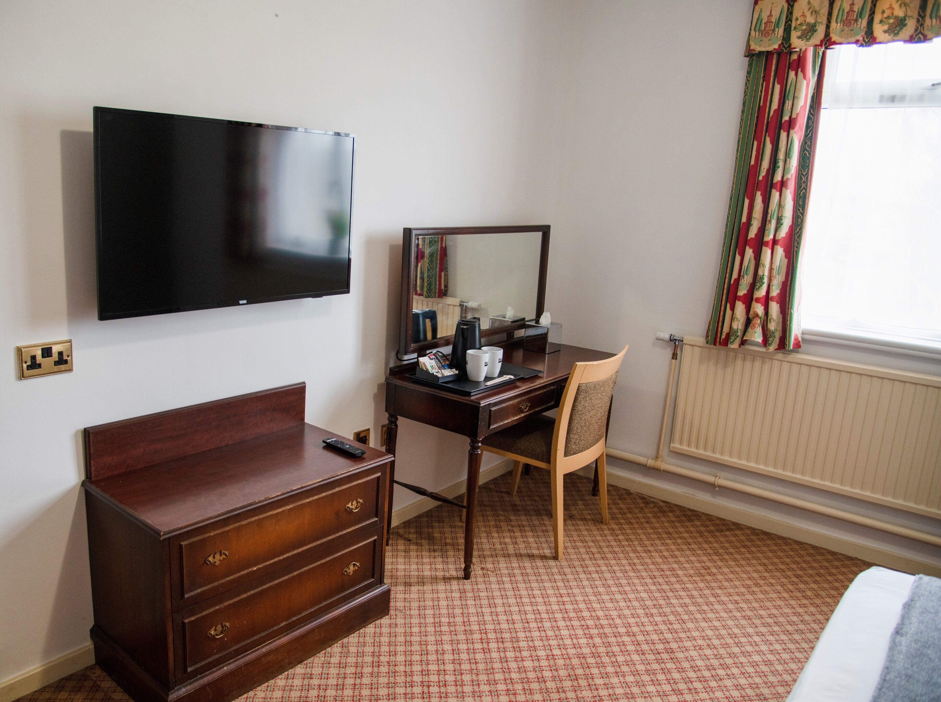 A hotel room with a flat-screen TV mounted on the wall, a wooden dresser with drawers, a desk with a chair, and a window with patterned curtains.