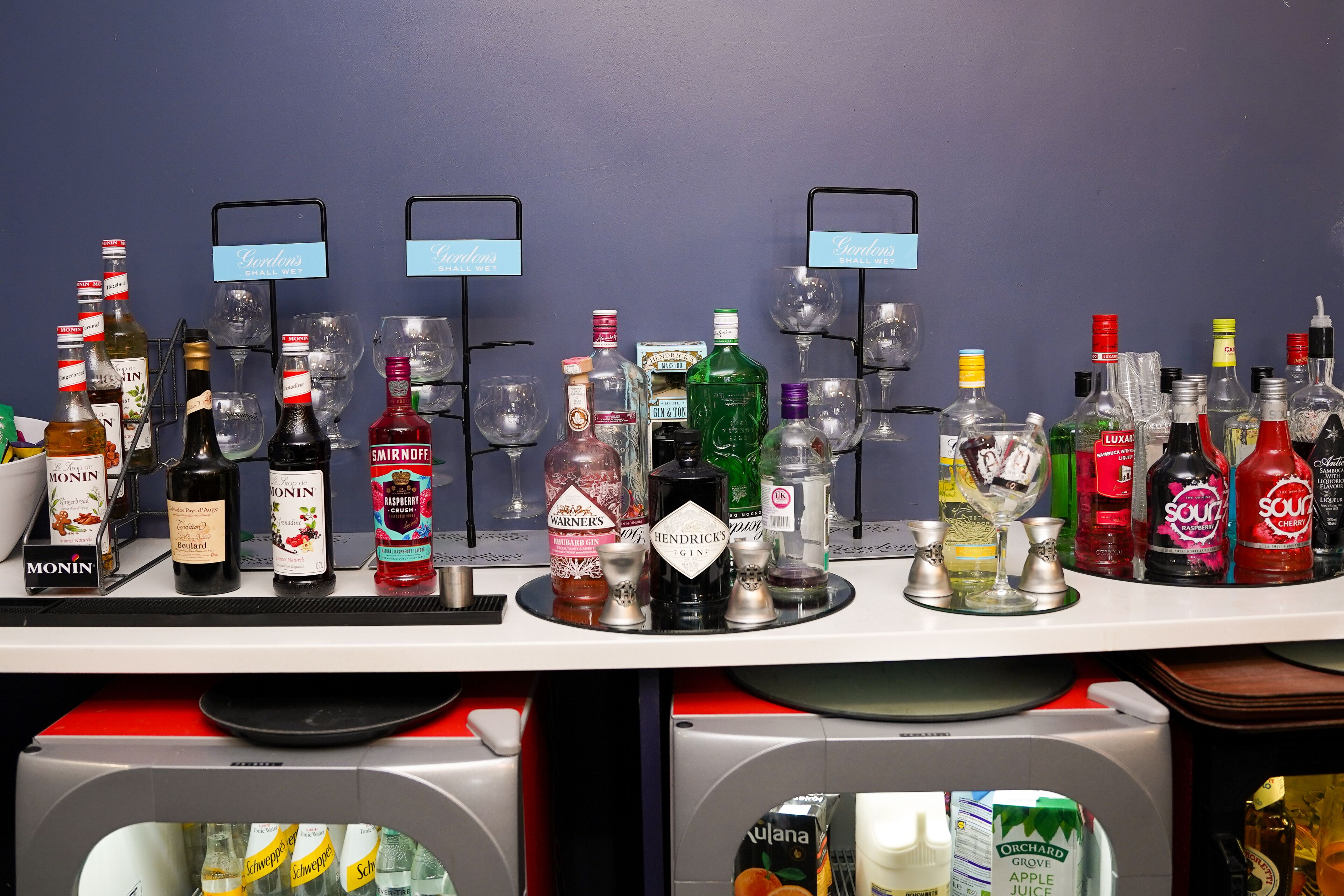 Een bar met verschillende drankflessen en glazen, waaronder merken als Monin, Smirnoff, Hendrick's en LUXA.