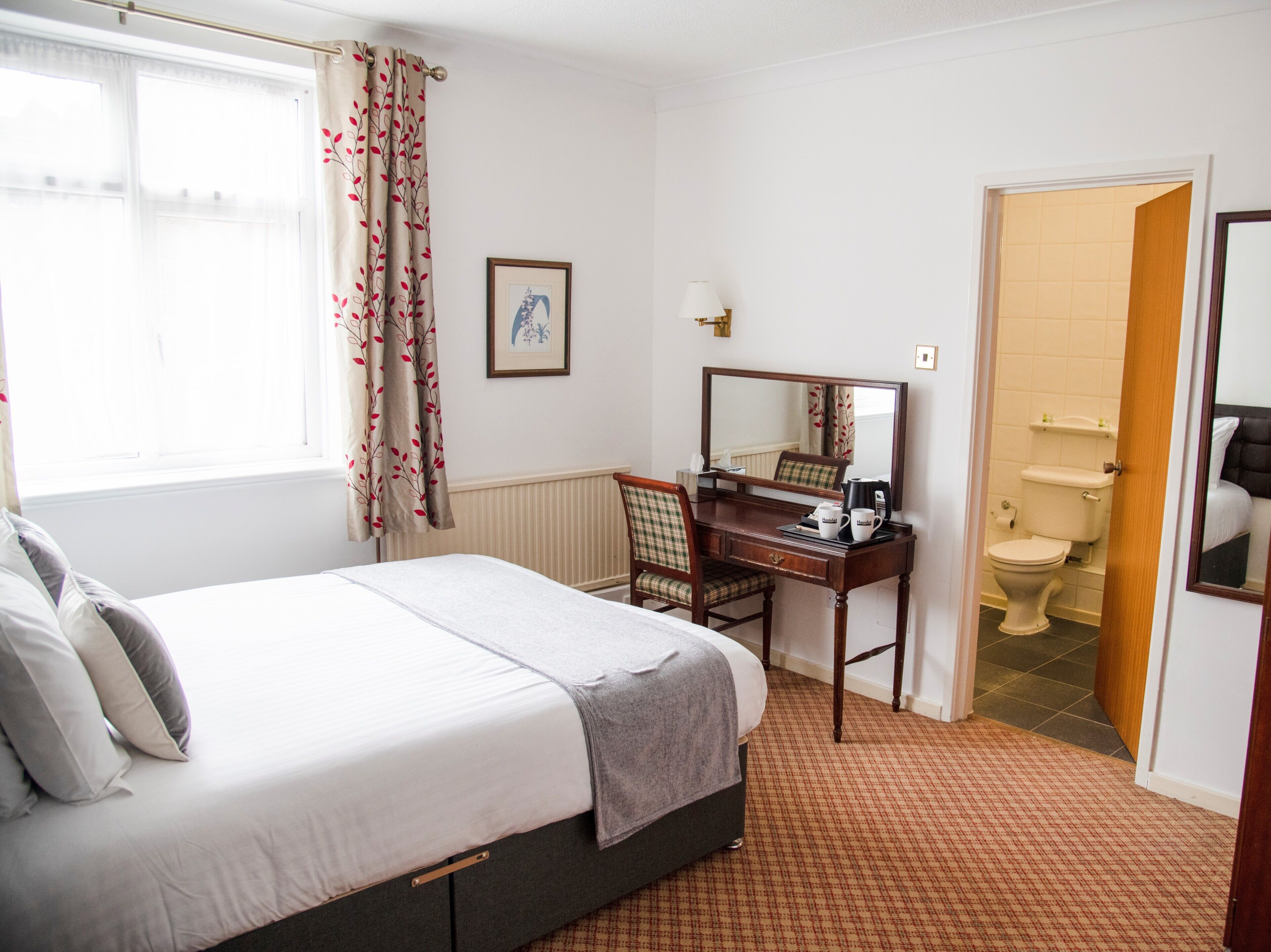 Quarto de hotel com cama, escrivaninha, cadeira, espelho, janela com cortinas, quadro pendurado na parede e porta do banheiro.