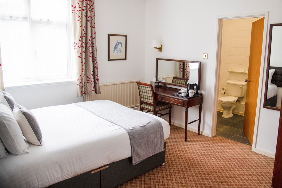 A hotel room with a bed, a desk with a chair, a mirror, a framed picture on the wall, and a bathroom visible through an open door.