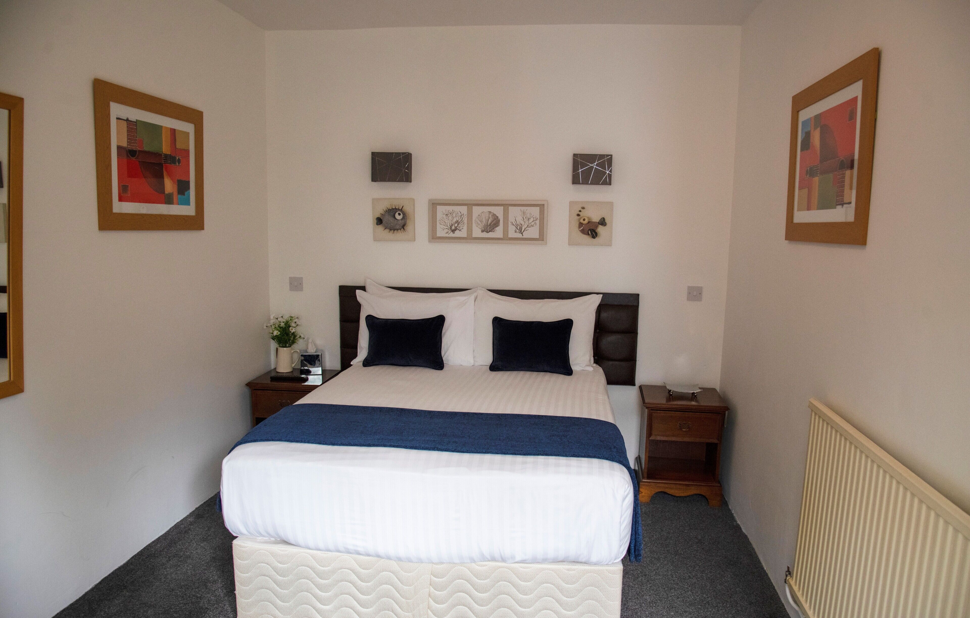 Una cama bien hecha con un edredón azul, dos mesitas de noche con lámparas, y tres cuadros en la pared.