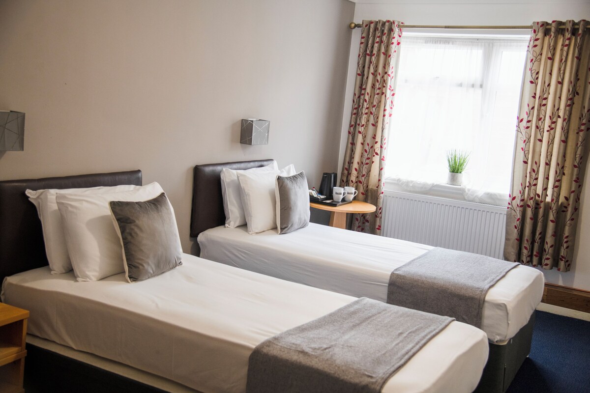A hotel room with two single beds, a small table with a lamp, a window with curtains, and a radiator.