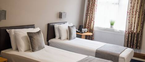 A hotel room with two single beds, a small table, a radiator, and curtains with a leaf pattern.