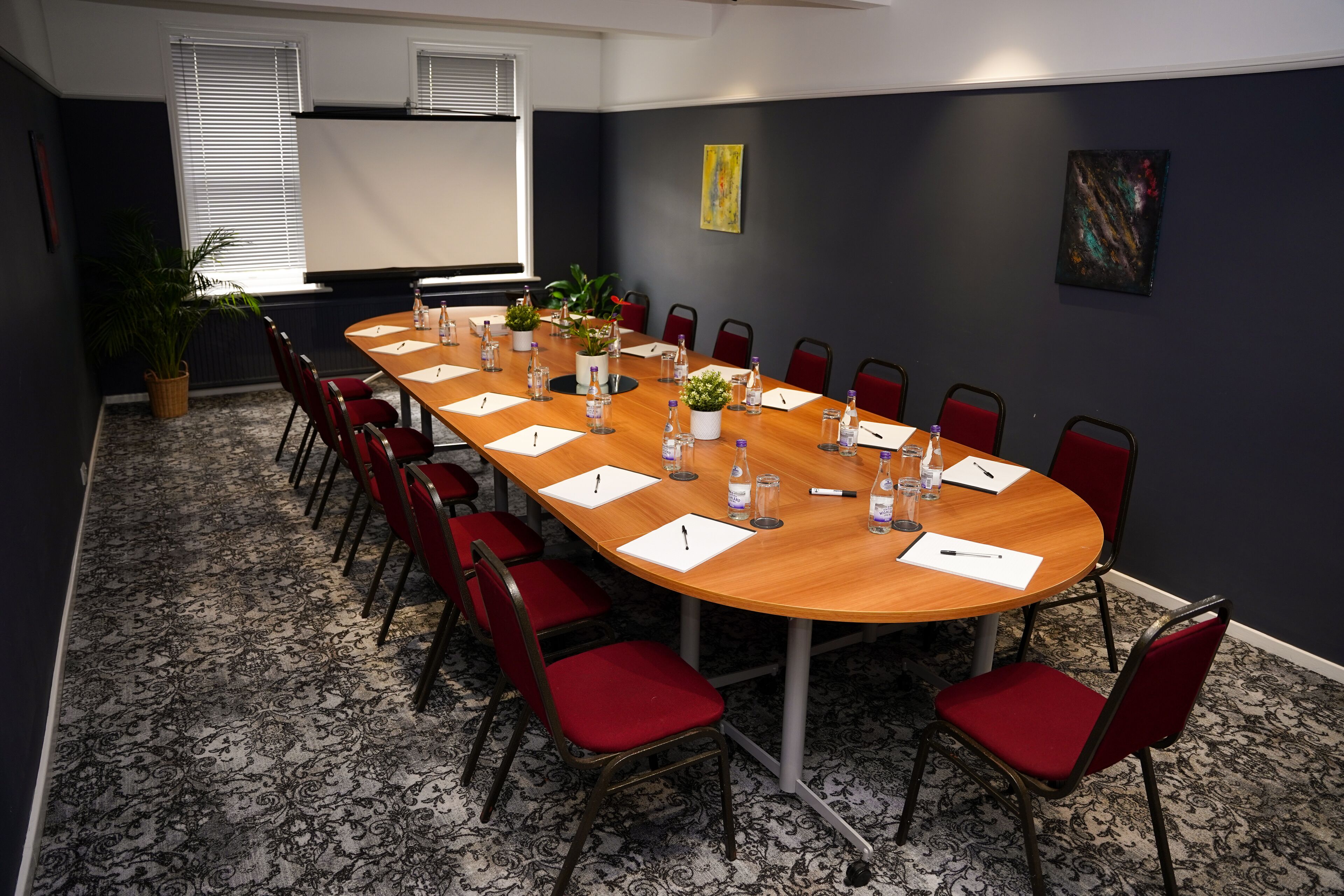 A conference room with a long table, red chairs, and a projector screen.