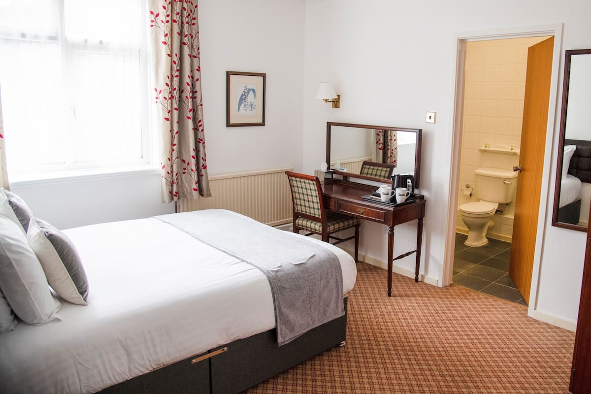 A hotel room with a bed, a desk with a chair, a mirror, a framed picture on the wall, and a bathroom visible through an open door.