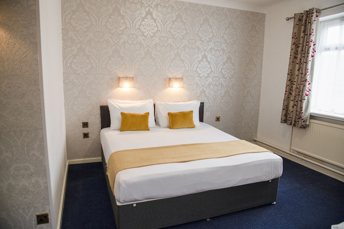A hotel room with a bed, two wall-mounted lights, a window with curtains, and patterned wallpaper.