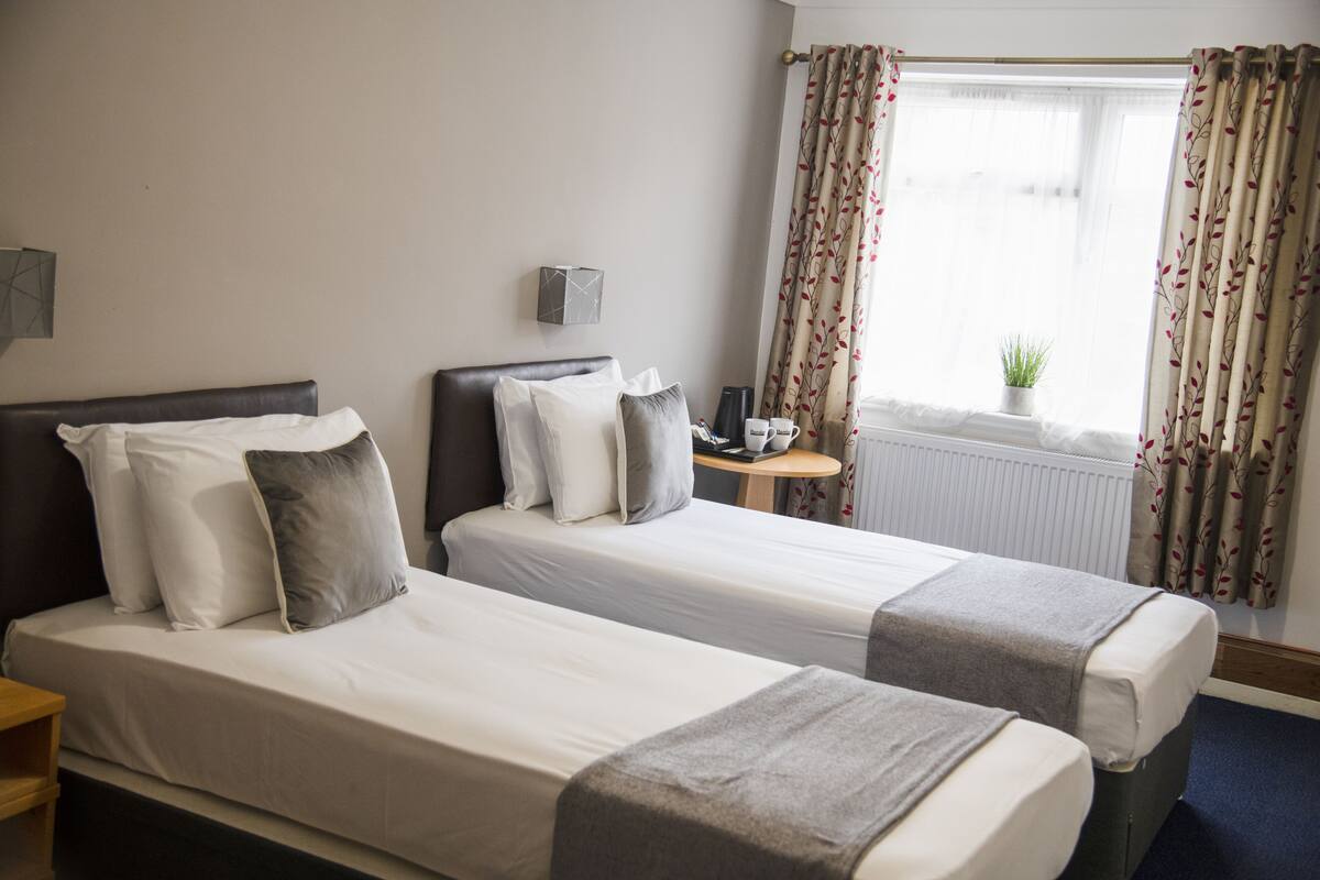 A hotel room with two single beds, a small table with a lamp, a window with curtains, and a radiator.