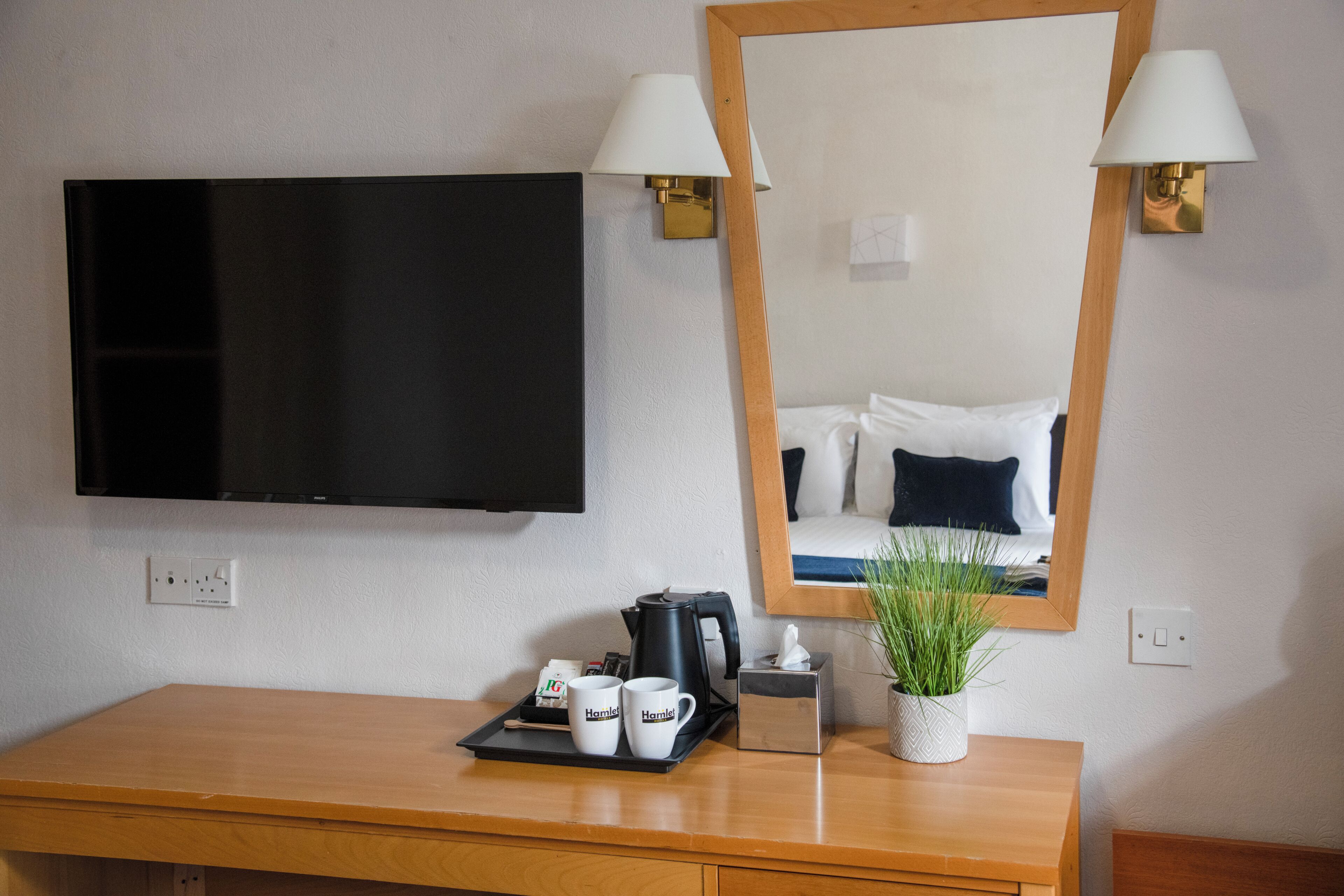 Quarto de hotel com televisão de tela plana, escrivaninha de madeira com bandeja de canecas, cafeteira e planta em vaso.