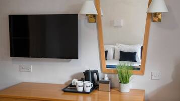 Quarto de hotel com televisão de tela plana, escrivaninha de madeira com bandeja de canecas, cafeteira e planta em vaso.