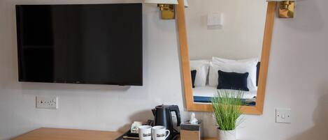 A hotel room with a flat-screen TV, a wooden desk with a tray of mugs, a coffee maker, and a potted plant.
