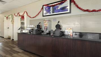 A hotel reception area with a dark wood counter, a waffle maker, a coffee machine, and a framed picture on the wall.