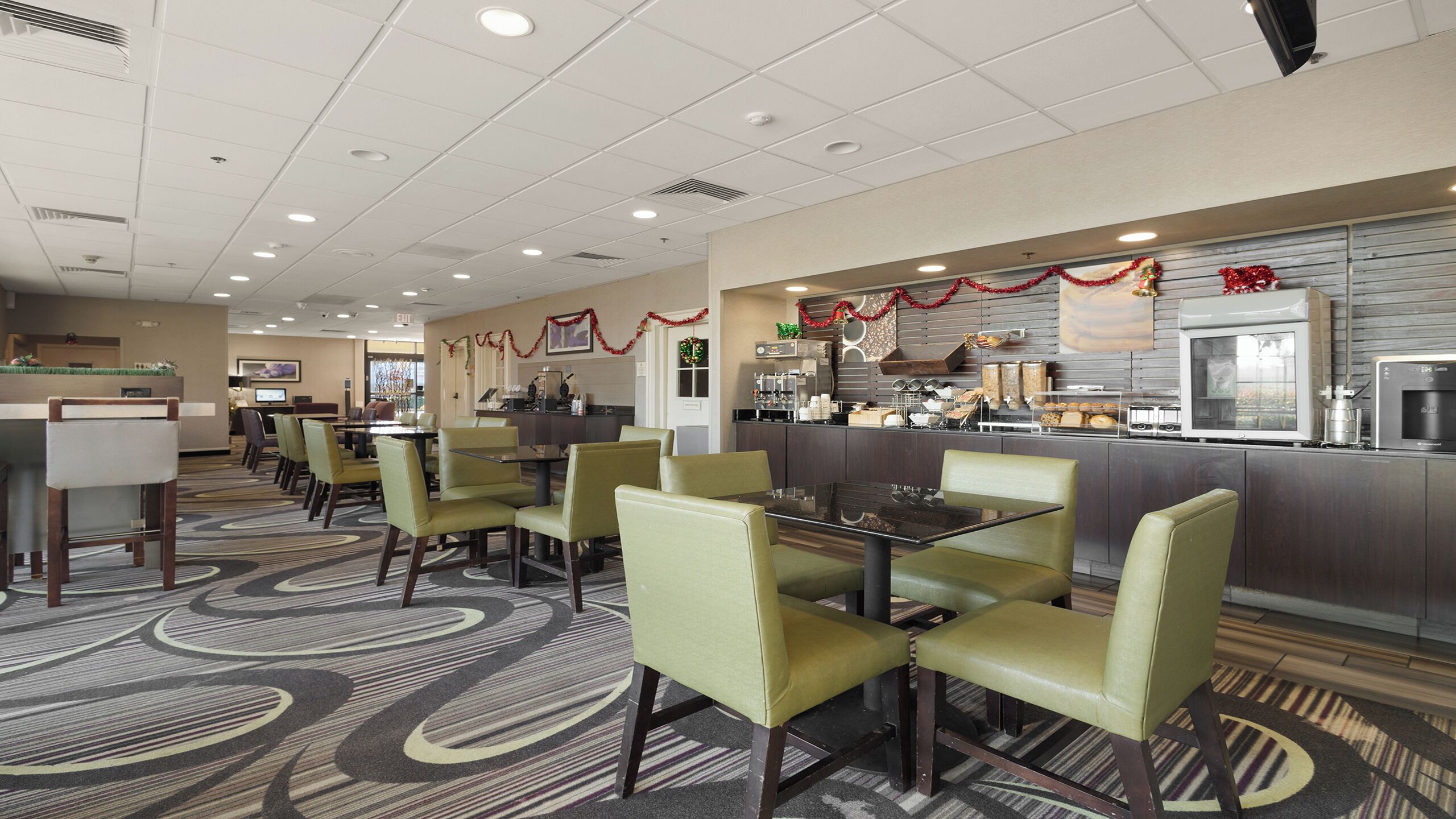 A hotel dining area with multiple tables and chairs, a kitchen area with appliances, and holiday decorations.