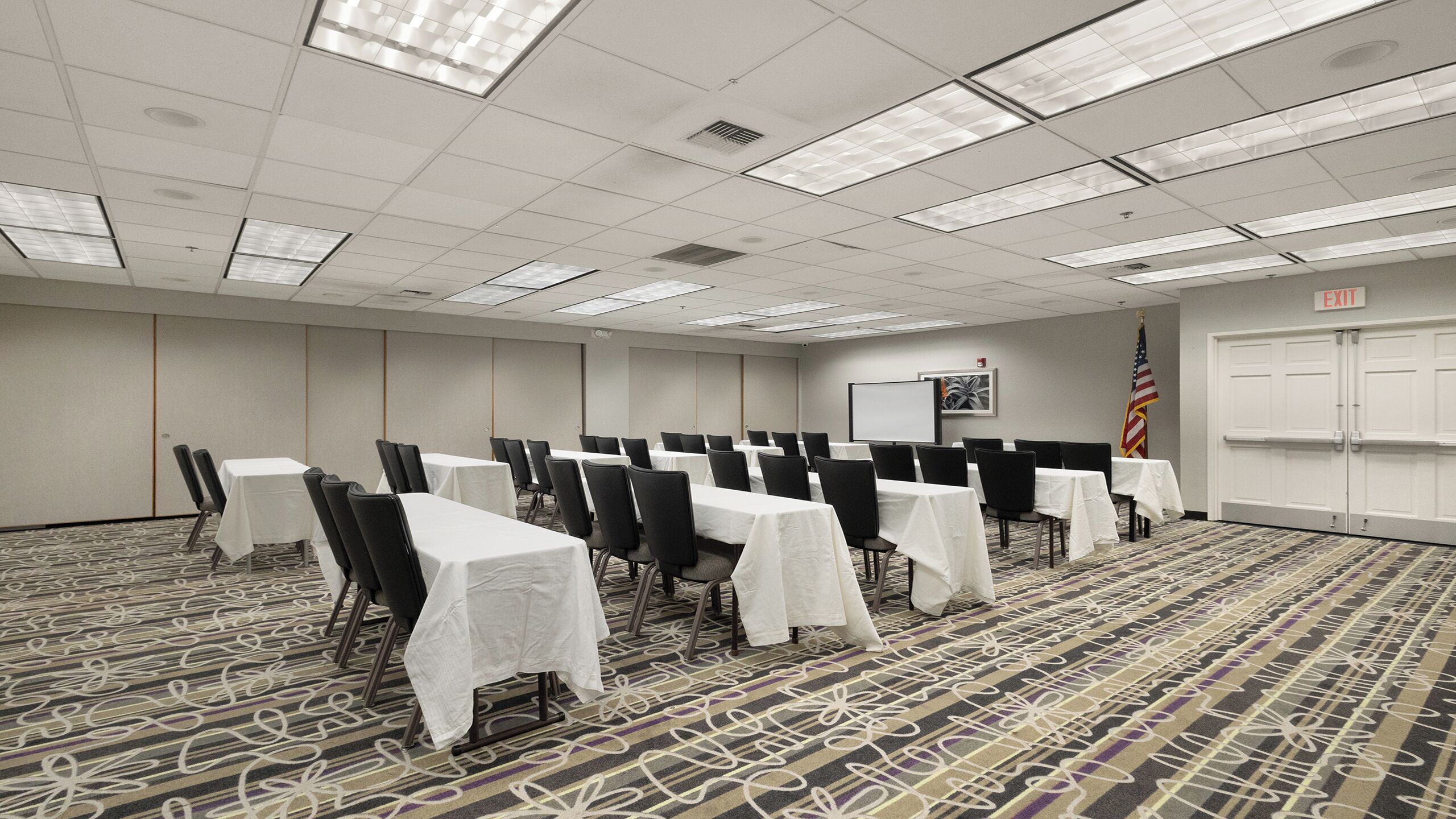 A conference room with rows of tables and chairs, a projector screen, an American flag, and an exit sign.