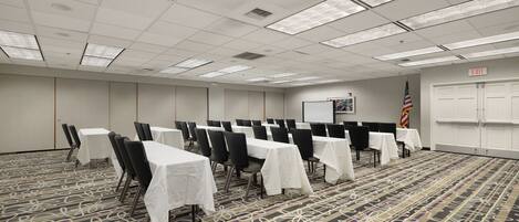 A conference room with rows of tables and chairs, a projector screen, an American flag, and an exit sign.