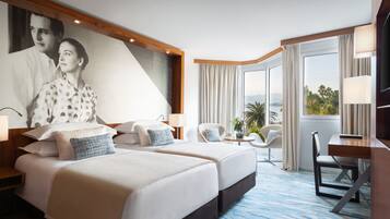 A hotel room with two beds, a desk, a chair, a TV, and a large window with curtains.
