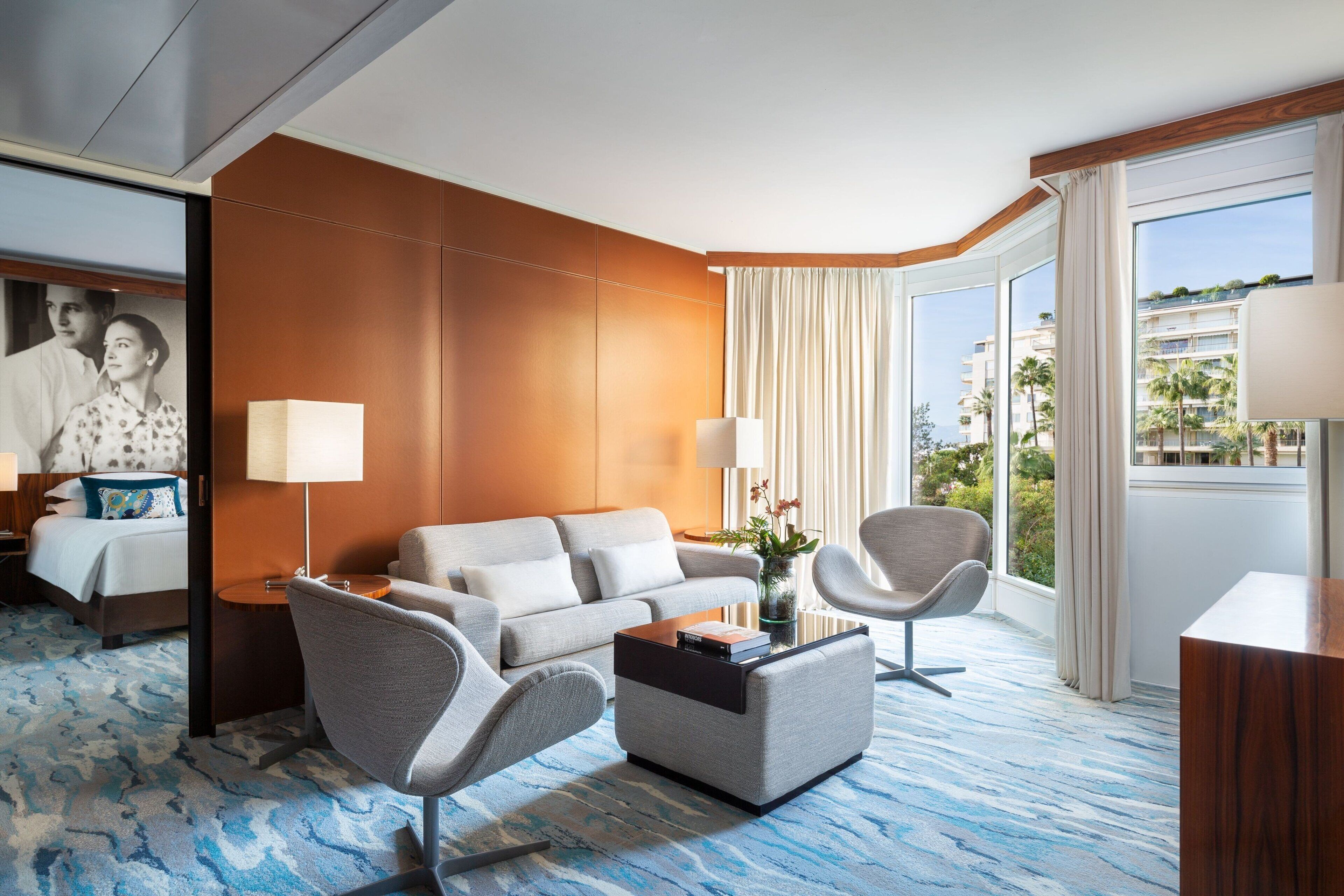 A hotel room with a sofa, two armchairs, a coffee table, a lamp, and a view of palm trees and buildings.
