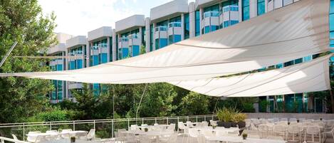 An outdoor patio with white tables and chairs, a large white canopy, and a modern building with blue windows in the background.