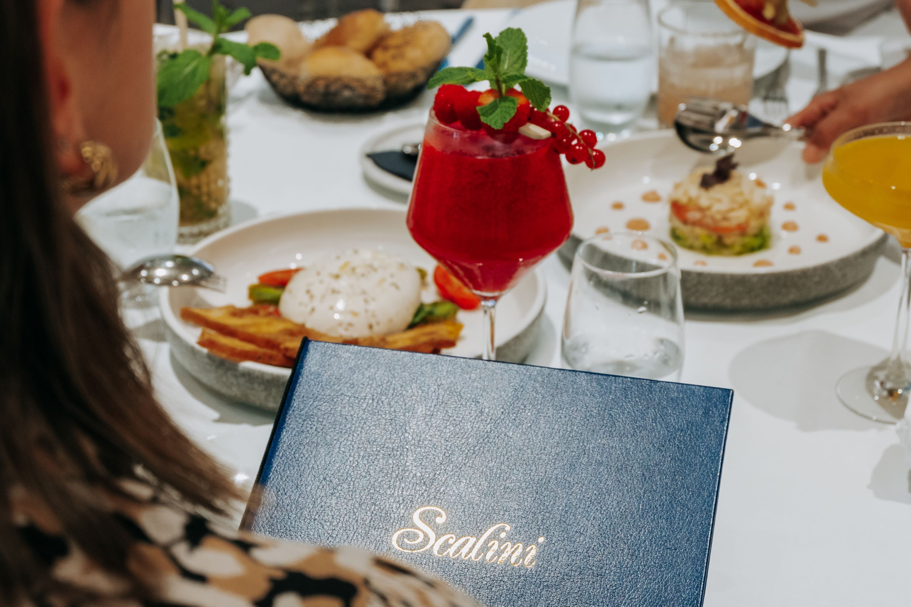 Une table à manger avec différents plats, une boisson rouge aux fruits des bois et un menu intitulé « Scalini ».