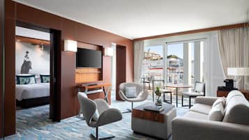 A hotel room with a bed, a TV, a seating area with a sofa and chairs, and a view of the city through large windows.