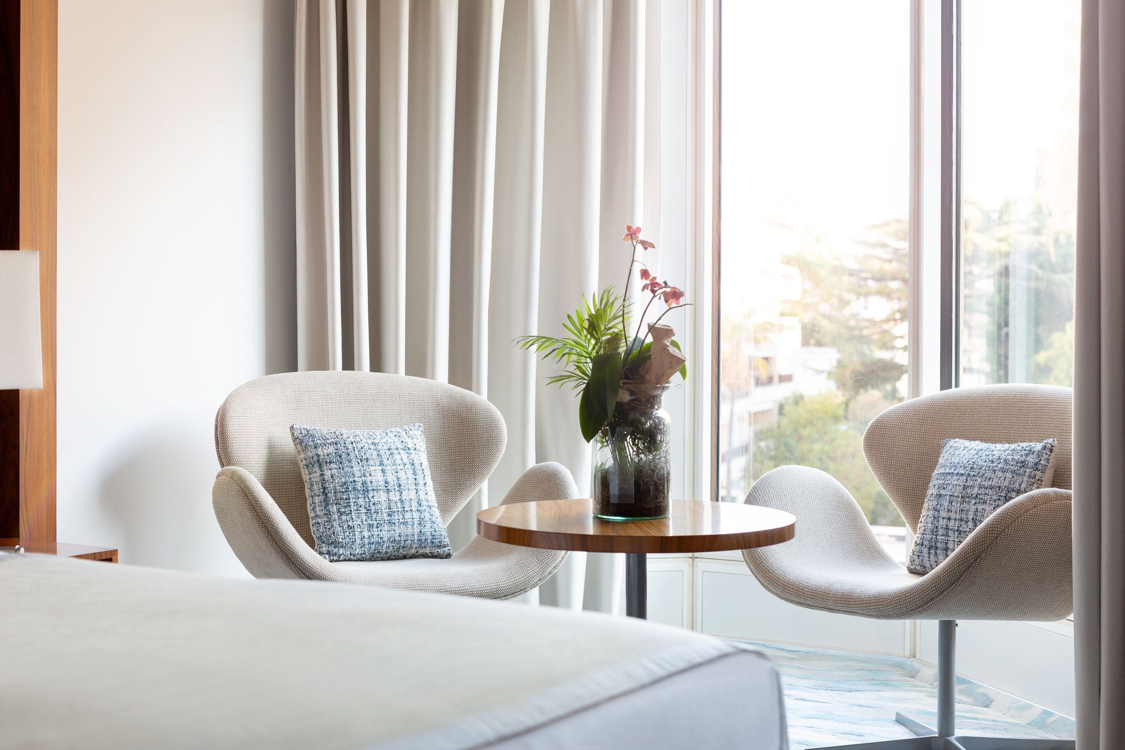 Ein Hotelzimmer mit einem Bett, zwei Sesseln, einem kleinen runden Tisch und einer Vase mit Blumen.